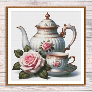 Könnte beinhalten: Eine weiße Porzellan-Teekanne mit goldenen Verzierungen und einem zarten Blumendesign. Die Teekanne wird von einer passenden Teetasse und einem Unterteller begleitet, die ebenfalls mit Blumendekoren verziert sind. Eine rosa Rose mit grünen Blättern befindet sich im Vordergrund.