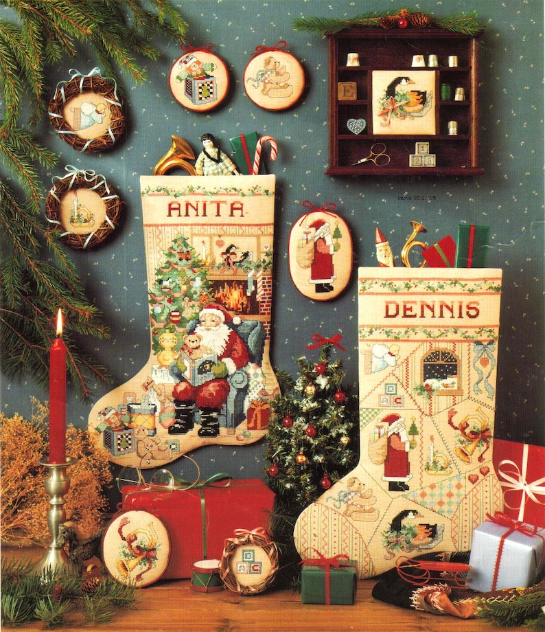May include: A festive Christmas scene featuring two embroidered stockings, one labeled "ANITA" and the other "DENNIS", filled with gifts and decorations. Ornaments, a small Christmas tree, and wrapped presents complete the holiday display.