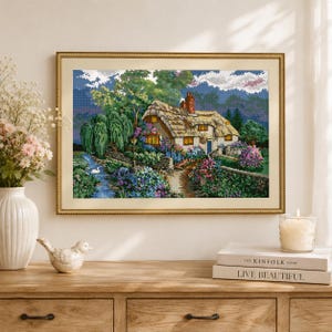 May include: A framed cross-stitch embroidery depicting a thatched-roof cottage with a brick chimney, surrounded by lush green trees, vibrant purple and blue flowers, a stone wall, a winding path, and a small stream with a white swan in the water.