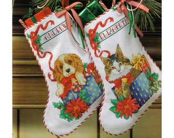 May include: Two white Christmas stockings with red and white trim. The stocking on the left has a puppy peeking out of a blue and white gift box with a red poinsettia and green holly. The stocking on the right has a cat peeking out of a blue and white gift box with a red poinsettia and green holly. The stocking on the left has the name "Andrew" embroidered on it. The stocking on the right has the name "Elizabeth" embroidered on it.
