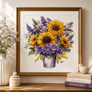 May include: A framed cross-stitch artwork depicting a bouquet of yellow sunflowers and purple flowers arranged in a light purple bucket. The floral arrangement features vibrant yellow petals with dark centres and clusters of purple blossoms and green leaves.