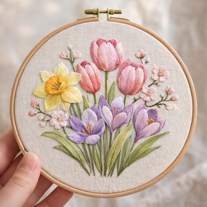 Puede incluir: Bordado floral en un aro de madera. El diseño presenta un ramo de flores primaverales con narcisos amarillos, tulipanes rosas y crocus morados sobre tela crema. El bordado está en un aro de madera redondo.