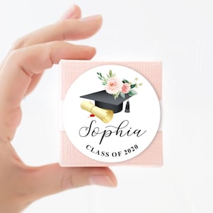 May include: A white round sticker with a black graduation cap, a gold scroll, and pink flowers. The text "Sophia Class of 2020" is printed on the sticker.