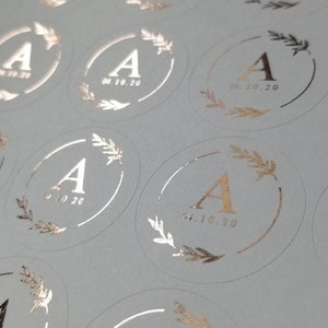 May include: A sheet of white paper with multiple round stickers. Each sticker has a gold foil design of a wreath with the letter A in the center and the date 04.10.20 printed below.