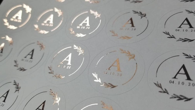 May include: A sheet of white paper with multiple round stickers. Each sticker has a gold foil design of a wreath with the letter A in the center and the date 04.10.20 printed below.