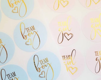 May include: A close-up of several round stickers with gold foil lettering. The stickers are on a white background. The stickers say "Team Boy" and "Team Girl" with a heart symbol.