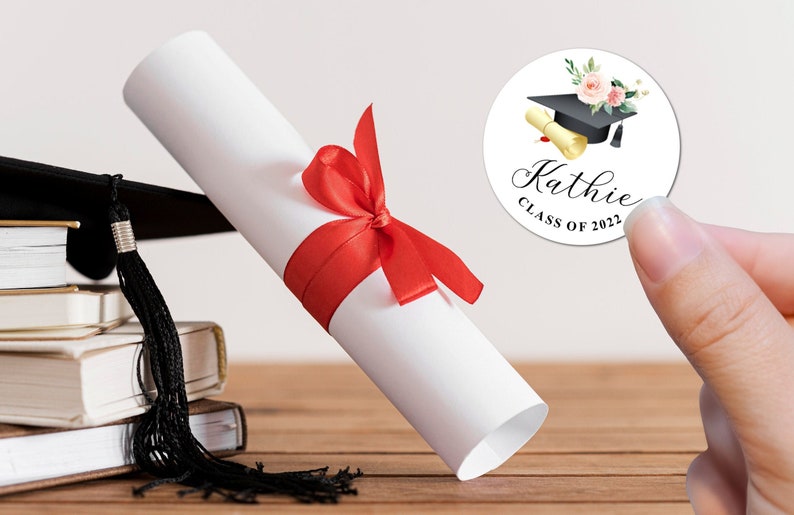 May include: A white round sticker with a black graduation cap, a gold scroll, and pink flowers. The text on the sticker reads "Kathie Class of 2022".
