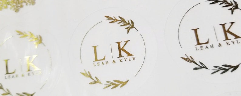 May include: Gold foil stickers with a simple design featuring the initials "LK" surrounded by a wreath of leaves. The text "Leah & Kyle" is written below the initials.