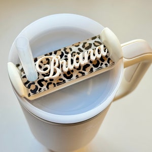 May include: White travel mug with a white lid and a handle. The lid has a clear straw holder and a leopard print sticker with the name "Briana" on it.