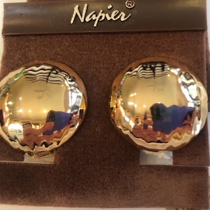 Puede incluir: Un par de pendientes de clip dorados y texturizados. Los pendientes son redondos con un efecto de metal martillado, que refleja la luz. Se exhiben en una tarjeta de fieltro marrón con el nombre de la marca "Napier" en letras plateadas.