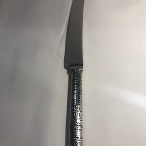 May include: A silver butter knife with a patterned handle. The handle is decorated with a repeating geometric design.