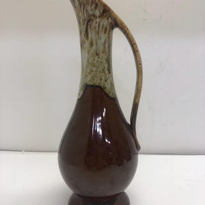 May include: A tall ceramic pitcher with a bulbous base and a slender, elongated neck. The lower portion features a glossy dark brown glaze, while the upper neck and handle transition into a textured, mottled cream and light brown drip glaze finish.
