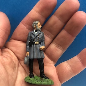 May include: A miniature figurine of a man in a gray military uniform, holding a hat. The figurine stands on a green base, with black boots and pants. The man has a gray beard and is looking to the side. The uniform has gold accents.