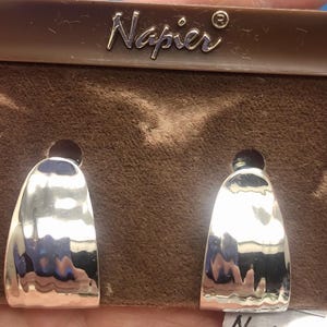 Puede incluir: Un par de pendientes de aro plateados con textura martillada, presentados en una tarjeta marrón. Los pendientes tienen un diseño curvo y abierto, bajo la marca "Napier".