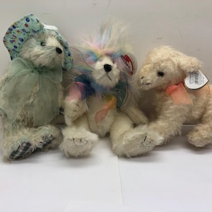 Könnte beinhalten: Drei Plüschtiere werden gezeigt. Ein Teddybär mit Blumenhut und grünem Band, ein Löwe mit Regenbogenhaar und farbigem Band und ein Lamm mit rosa Band. Alle haben Anhänger.