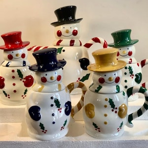 May include: A collection of snowman-shaped teapots with colorful top hats and candy cane handles. The teapots are white with red, green, and yellow accents, including holly and berry designs. Each teapot has a smiling face and is designed for holiday use.
