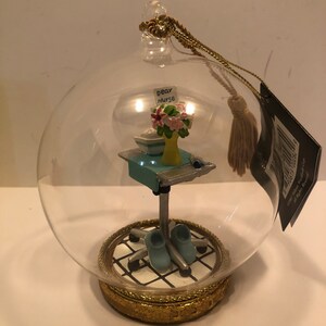 Peut inclure: Une boule de Noël en verre avec une scène miniature à l'intérieur. Un petit bureau avec une note qui dit "Dear Nurse" et un bouquet de fleurs se trouve sur une chaise roulante. La boule est décorée d'une bordure dorée.