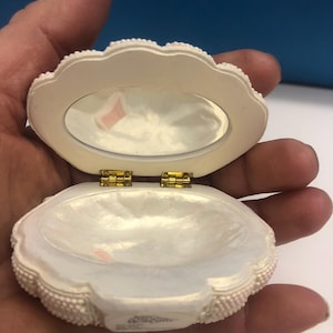 May include: Open, off-white clam shell-shaped trinket box with a small mirror inside the lid. The box is decorated with small, raised beads around the edges and has gold-coloured hinges. The interior is pearlescent.