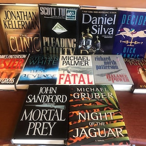 Può includere: Una collezione di libri con copertina rigida con vari titoli e autori, tra cui "The Clinic" di Jonathan Kellerman, "Mortal Prey" di John Sandford e "Night of the Jaguar" di Michael Gruber. I libri sono disposti su una superficie di legno.