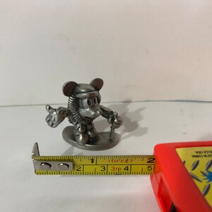 以下が含まれることがあります： 冬服を着てスキーの棒を持ったミッキーマウスの銀色のピューター製フィギュア。台座の上に立っています。フィギュアの高さは約5センチメートルです。