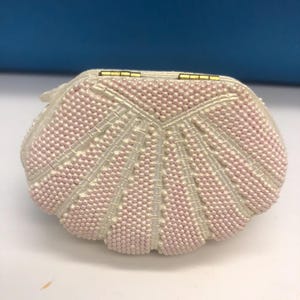 May include: A small, shell-shaped beaded purse. The purse is mainly off-white with a pattern of pink beads. The clasp is gold-coloured. The purse is likely vintage and suitable for special occasions.
