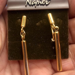 Puede incluir: Pendientes dorados con diseño de barra rectangular. Los pendientes cuelgan de una pequeña cadena y están sujetos a un poste dorado. Los pendientes se exhiben en una tarjeta marrón con el nombre de la marca "Napier" en letras plateadas.
