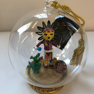 May include: A glass ornament featuring a hand-painted Native American figure with a headdress, holding a blue object. The figure is standing on a gold base with a cactus and a small pottery pot. The ornament is decorated with gold glitter.