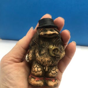 以下が含まれることがあります： 茶色の毛皮を着て、黒いシルクハットをかぶり、金の角笛を持っている生き物の気まぐれな置物。 この置物は、足に赤いアクセントが付いた詳細なデザインです。 背景は無地の青色です。