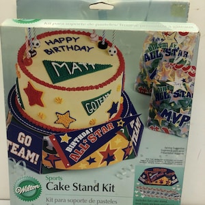 May include: A Wilton Sports Cake Stand Kit, featuring a decorated cake with "Happy Birthday" and "MAT" text. The kit includes a cake stand, treat bags, stickers, and more. The cake has a red, yellow, and green color scheme.