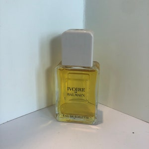 May include: A clear glass bottle of Ivoire de Balmain Eau de Toilette. The bottle has a white cap and a yellow label with black text.