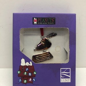 May include: A purple box with a silver Peanuts ornament inside. The ornament is a depiction of Snoopy, the beagle from the Peanuts comic strip. The box also features a Peanuts logo and the Lunt logo.