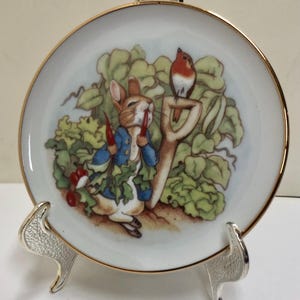 May include: A round white porcelain decorative plate with a gold-trimmed edge featuring a colorful illustration of a brown rabbit wearing a blue jacket holding red carrots in a garden with a small bird perched on a garden tool handle.