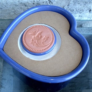 May include: A blue heart-shaped ceramic dish with a brown ceramic tile featuring a floral design in the center.