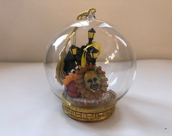 Puede incluir: Un adorno de vidrio con base dorada y una escena en el interior de una cara de payaso con un diseño de sol amarillo y naranja, figuras negras y borlas amarillas.