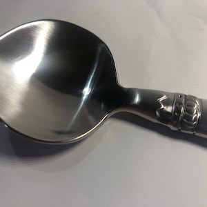 May include: A silver spoon with a rounded bowl and a detailed handle. The handle has a crown design and a raised inscription.