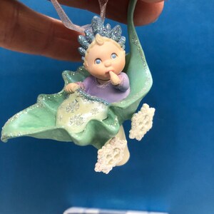 May include: A baby ornament with a light blue crown and a purple shirt, nestled in a green leaf-shaped cradle. The ornament has glitter accents and snowflake details. A pink ribbon is attached for hanging.