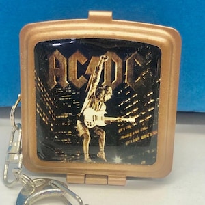 May include: A gold-coloured, rectangular keychain featuring an image of the band AC/DC. The image shows a guitarist playing in front of a cityscape. The keychain has a hinged design and a silver-coloured clasp.