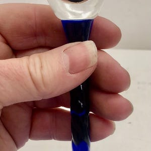 Puede incluir: Una copa de vino transparente con base azul y tallo retorcido azul oscuro y azul cobalto. La copa est&aacute; sujeta por una mano. La base tiene aproximadamente 7,6 cm de di&aacute;metro.