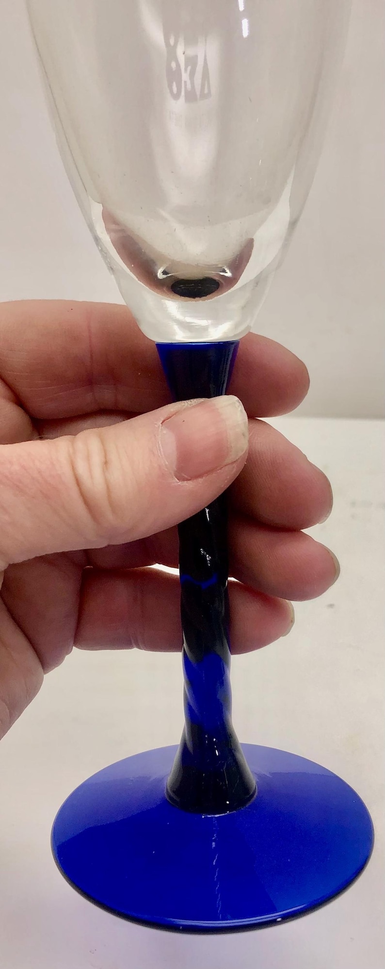 Puede incluir: Una copa de vino transparente con base azul y tallo retorcido azul oscuro y azul cobalto. La copa est&aacute; sujeta por una mano. La base tiene aproximadamente 7,6 cm de di&aacute;metro.