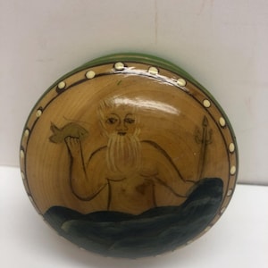 Op de afbeelding: Een ronde, houten decoratieve plaat met een geschilderd beeld van een bebaarde figuur die een vis en een drietand vasthoudt. De figuur bevindt zich in een zee van donkerblauwe golven. De plaat heeft een groene rand en een rand van witte stippen.