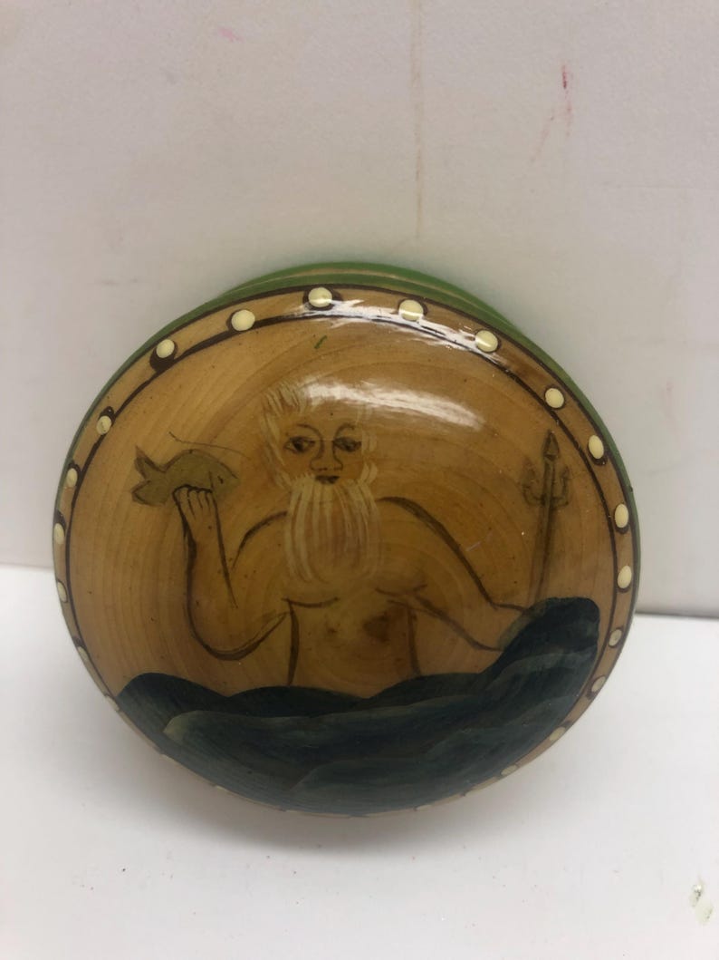 Op de afbeelding: Een ronde, houten decoratieve plaat met een geschilderd beeld van een bebaarde figuur die een vis en een drietand vasthoudt. De figuur bevindt zich in een zee van donkerblauwe golven. De plaat heeft een groene rand en een rand van witte stippen.