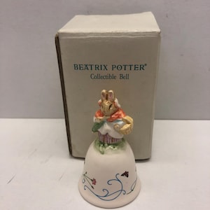 Puede incluir: Una campana de cerámica blanca con una figurita de Peter Rabbit de Beatrix Potter en la parte superior. La campana está decorada con diseños florales azules y verdes y una mariposa. La caja dice "Beatrix Potter Collectible Bell".
