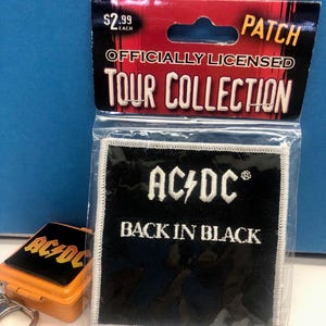 May include: A black and white AC/DC "Back in Black" patch from the Tour Collection. The patch is square with white stitching around the edges. The packaging includes the text "OFFICIALLY LICENSED". An AC/DC keychain is also visible.