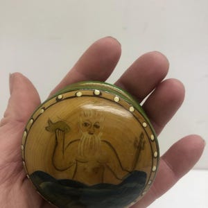 Op de afbeelding: Ronde, handgeschilderde houten doos met een deksel. De doos toont een afbeelding van een bebaarde figuur die een vis en een drietand vasthoudt, tegen een gele achtergrond met blauwe golven. De deksel is afgezet met groene en witte stippen.