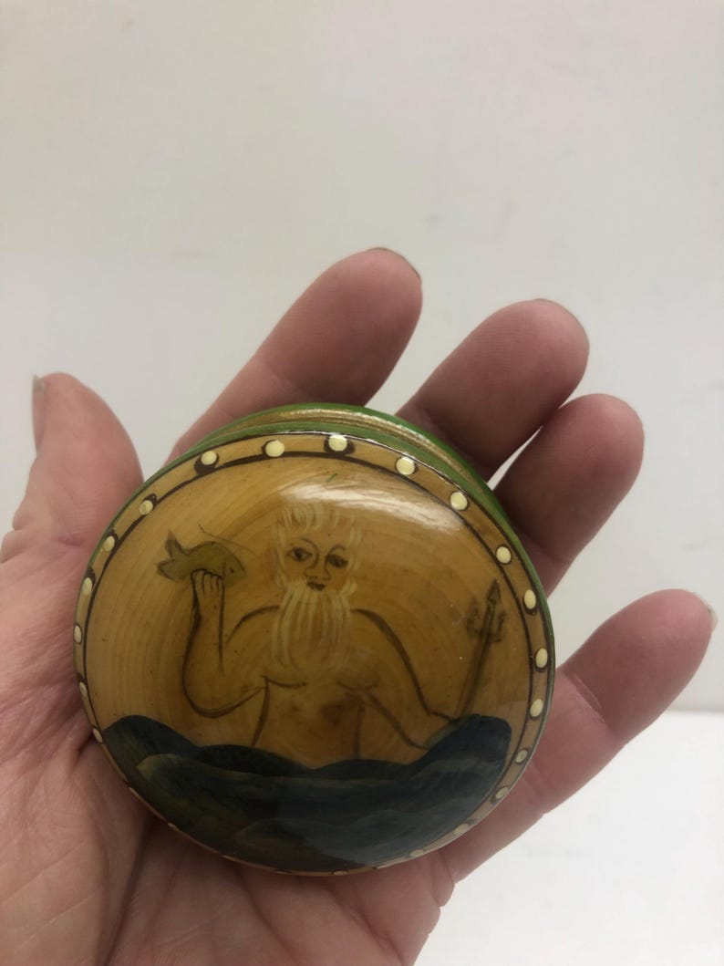 Op de afbeelding: Ronde, handgeschilderde houten doos met een deksel. De doos toont een afbeelding van een bebaarde figuur die een vis en een drietand vasthoudt, tegen een gele achtergrond met blauwe golven. De deksel is afgezet met groene en witte stippen.