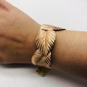 May include: Gold-toned leaf-shaped bracelet. The bracelet is made of metal and has a textured surface. The bracelet is on a person's wrist.