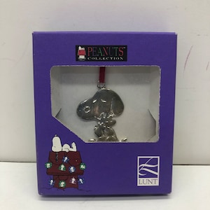 May include: A purple box with a silver Peanuts ornament inside. The ornament is of Snoopy holding a Christmas tree. The box has the Peanuts Collection logo and the Lunt logo.
