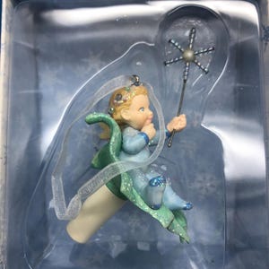 May include: A Christmas ornament featuring a seated cherub in a light blue outfit with glitter accents. The cherub holds a silver wand with a starburst design. The ornament has a white base and green and blue wings. The ornament is in a clear plastic box.