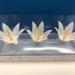 May include: Three white lily flower decorations with yellow centers are displayed in a clear plastic container. The container sits on a base with a snowflake pattern. The background is a solid blue.
