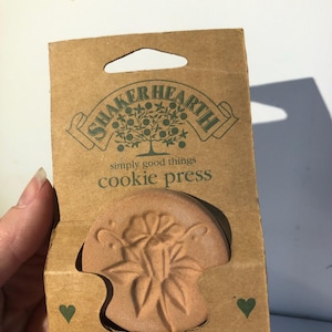 May include: A round, light brown ceramic cookie press featuring a floral design, packaged in a tan cardboard sleeve. The packaging displays the text SHAKER HEARTH, simply good things, and cookie press, with small green heart icons on the bottom corners.